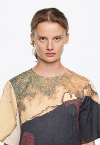 Top de manga corta hecho de una tela texturizada con una mezcla de colores terrenales: beige, verde y gris oscuro con patrones abstractos.