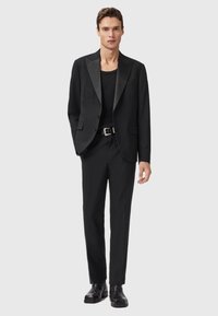 Completo nero con rever in raso, chiusura a un bottone e pantaloni slim fit abbinati a un top in tinta e stivali alla caviglia. Texture liscia.