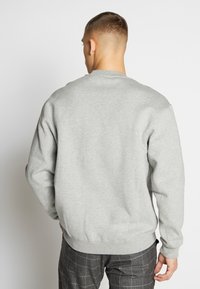 Šedá mikina s kulatým výstřihem, dlouhými rukávy a žebrovanými manžetami. Měkká tkanina a volný střih. Nosič má na sobě kostkované kalhoty.