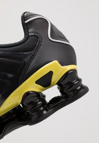 Talons de chaussure de sport noirs et jaunes avec amorti d'air courbé et dessus synthétique noir en mesh et perforé sur fond clair.