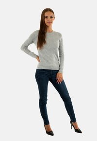Maglione grigio a maniche lunghe realizzato in un tessuto morbido, abbinato a jeans slim-fit blu scuro e a scarpe con tacco nere. Il modello posa in posizione eretta.