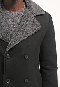 Svart ullkappa med en grå, luddig krage och dubbelknäppt knäppning. Strukturerad tyg med två framfickor.