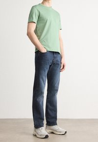 Lys grøn bomulds t-shirt med rund hals, parret med mørkeblå denim jeans og grå træningssko. Står foran en neutral baggrund.