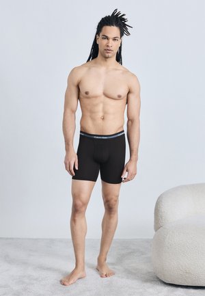 Homme aux longues dreadlocks portant un boxer noir Calvin Klein, debout pieds nus sur un tapis à côté d'un fauteuil blanc texturé.