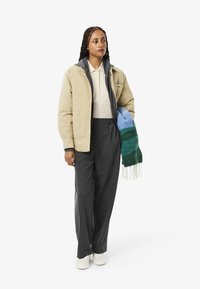 Chaqueta aislante beige sobre un sudadera gris, combinada con un polo beige y pantalones gris oscuro. Sosteniendo una bufanda multicolor y llevando unos zapatos blancos.