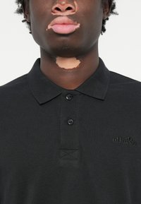 Fekete pólóing textúrált anyagból, gombos nyakkal, gallérral és hímzett logóval a mellkason.