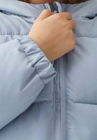 Dětská ruka svírá zip světle modré péřovky s elastickými manžetami, právě v akci zapínání.