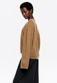 Jersey de punto beige con mangas largas, que presenta adornos negros distribuidos uniformemente por la tela. Textura suave, corte relajado.