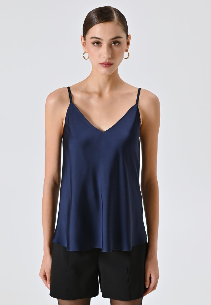 Donna con capelli pettinati all'indietro che indossa orecchini a cerchio dorati, una canottiera di raso blu navy e pantaloncini neri su uno sfondo chiaro e semplice.