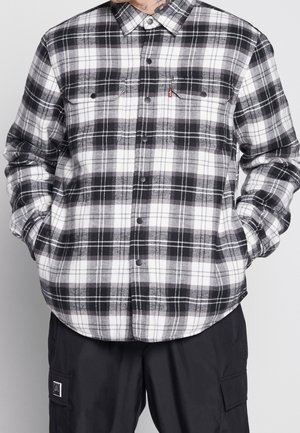 Personne portant une chemise à carreaux noire et blanche boutonnée avec poches poitrine et un pantalon cargo noir, debout les mains dans les poches.