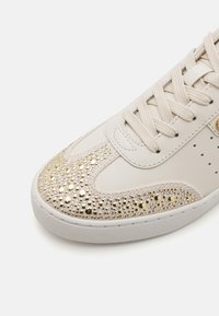 Beige sneaker met een glad leren bovenwerk, voorzien van een glinsterende gouden en zilveren studded neusstuk, en gestructureerde veters boven een witte zool.