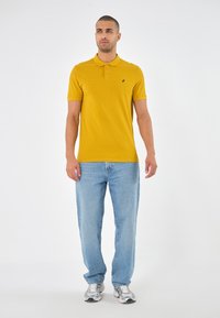 Polo shirt jaune moutarde à manches courtes, avec un rabat à trois boutons et un petit logo noir. Porté avec un jean en denim bleu clair et des baskets.