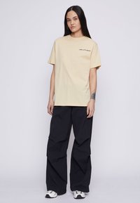 Lysegul bomull t-skjorte med korte ermer og svart tekst på brystet. Kombinert med løse, svarte, teksturerte bukser og sporty sko.