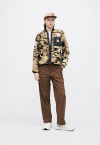 Cazadora bomber de forro polar en beige y negro con cremallera frontal y un bolsillo de parche negro. Pantalones marrones y zapatillas de color claro completan el look.