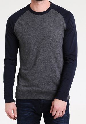 Hombre vistiendo una camiseta raglán de manga larga gris oscuro con mangas y cuello azul marino, combinada con vaqueros azul oscuro.