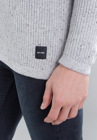 Lichtgrijze gebreide trui met een gestructureerde afwerking, voorzien van een zwart label met "ONLY & SONS" aan de onderzoom. Gecombineerd met een donkere spijkerbroek.