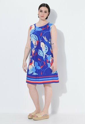 Kobieta stoi w niebieskiej sukience bez rękawów z czerwonym, beżowym i jasnoniebieskim kwiatowym wzorem oraz beżowych sandałach na platformie na białym tle.