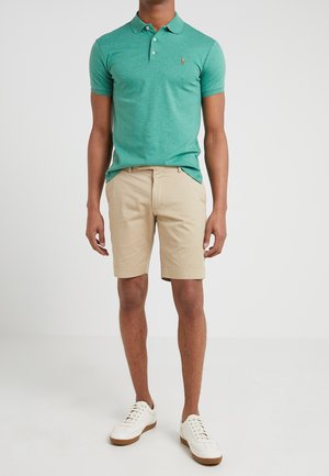 Grünes Poloshirt mit kurzen Ärmeln, kombiniert mit beigen Shorts. Weiße Sneakers mit braunen Sohlen. Einfaches Design und klassischer Schnitt.