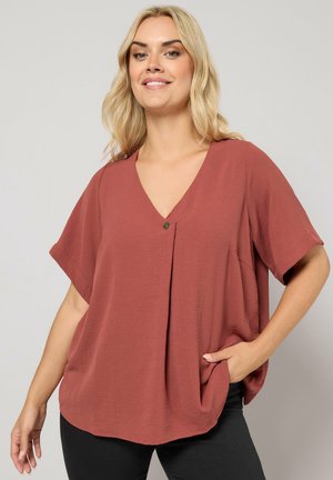 Smilende kvinde med blondt hår iført en løs, kortærmet bluse i rustfarve og sorte bukser, med den ene hånd i lommen.