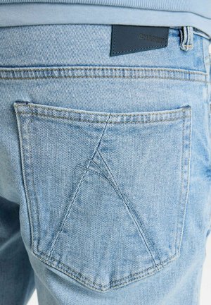 Svijetloplave traperice s istaknutom stražnjom džepom koja ima jedinstveni trokutasti uzorak šivanja i kožnu oznaku s natpisom "CHASING."