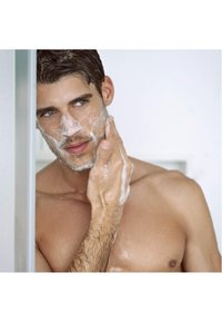 Un uomo con capelli castano chiaro e barba incolta applica un detergente schiumoso sul viso. Ha un fisico tonico e lo sfondo è luminoso e minimalista.