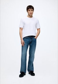 Junger Mann steht mit einer Hand in der Tasche, trägt ein schlichtes weißes T-Shirt, blaue Jeans, schwarzen Gürtel und schwarze Schuhe, vor weißem Hintergrund.