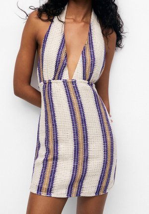 Mini-robe en maille sans manches avec un décolleté en V profond et des rayures verticales crème, violettes et sable, portée par une personne aux cheveux bouclés.