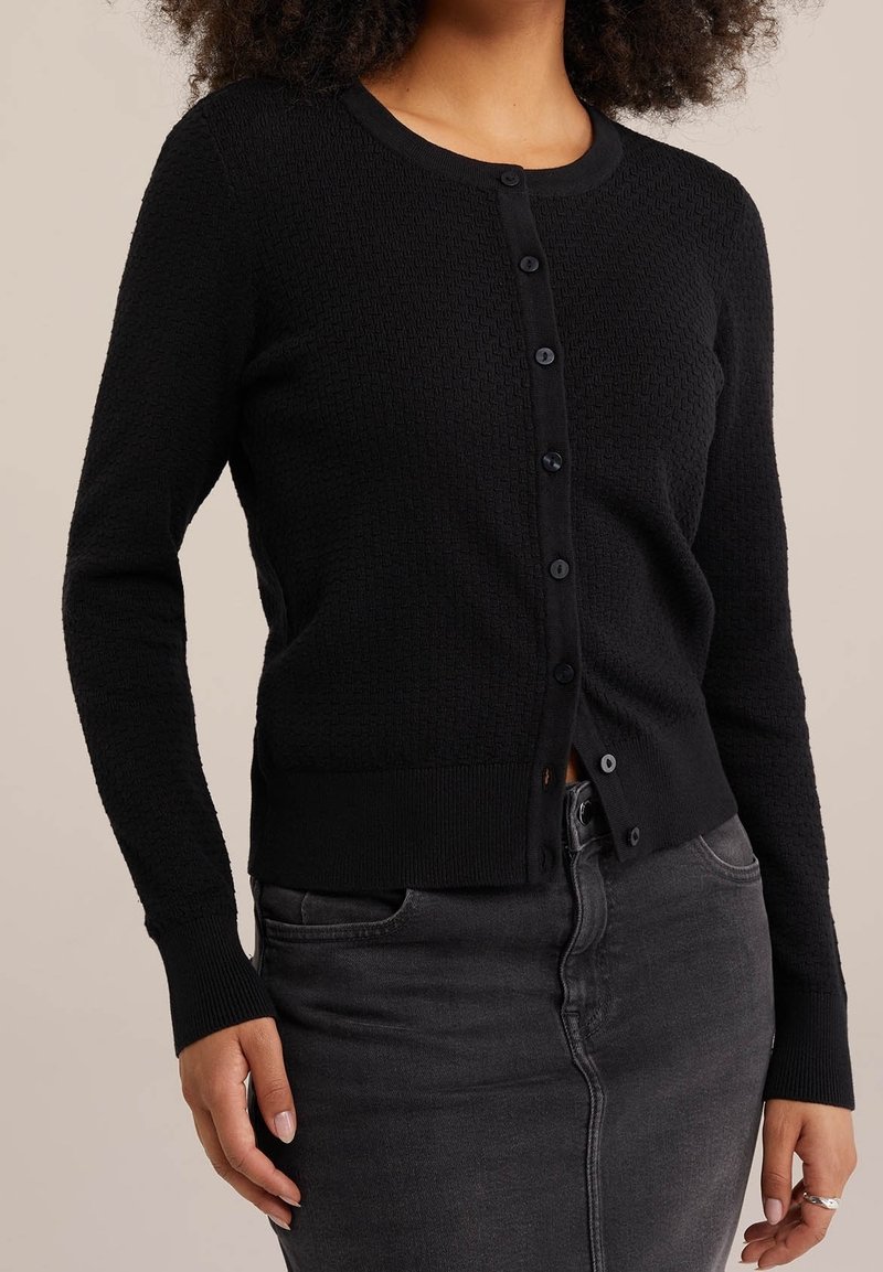 Svart texturerad kofta med rund halsringning, knappstängning och figursydda muddar, matchad med en grå kjol.