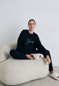 Frau mit dunklen Haaren und Creolen sitzt im Schneidersitz auf einem gebogenen beigen Sofa, trägt einen schwarzen Guess-Hoodie, Hosen und weiße Hausschuhe.