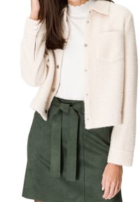 Mujer vestida con una chaqueta crema texturizada con botones, una blusa blanca con borde ondulado, y una falda verde de cintura alta con un cinturón atado.