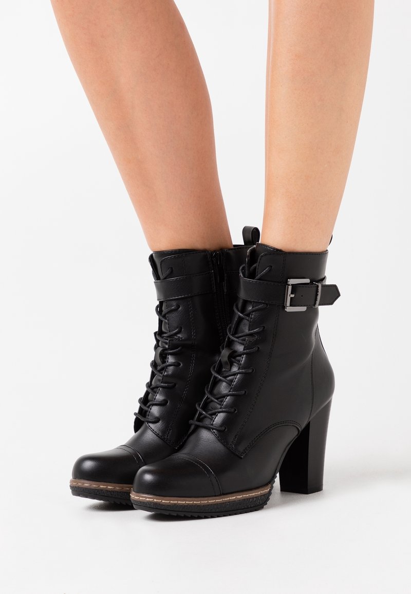 Bottines en cuir noir à talons hauts avec lacets, fermeture éclair latérale et sangle à boucle, portées sur des jambes nues sur fond blanc.