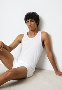 Top sin mangas y pantalones cortos de algodón blanco, con un diseño liso y forma ajustada. Tono de piel visible en brazos y piernas, con superficies texturizadas.