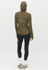 Felpa verde oliva con cappuccio e dietro allungato, abbinata a leggings neri con dettagli riflettenti e sneakers grigie con calzini bianchi.