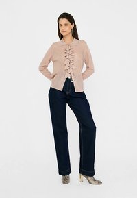 Žíhaný ružový sveter na gombíky s volánovým predným dielom, s golierom a zapínaním na gombíky, v kombinácii s tmavomodrými širokými džínsami a vzorovanými čižmami.