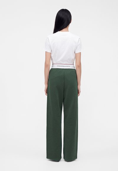 Femme aux longs cheveux noirs portant un haut blanc à manches courtes et un pantalon vert taille haute à jambes larges, debout de dos sur un fond blanc.