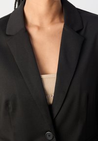 Schwarze Blazerjacke mit Reverskragen, Ein-Knopf-Verschluss und glatter Textur. Ein beiges Oberteil ist unter dem Blazer sichtbar.