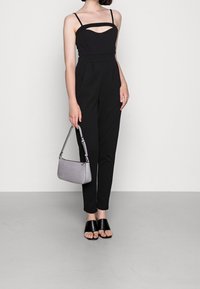 Svart jumpsuit med tunna axelband, utskärning vid bröstet, skräddarsydd passform, kombinerad med en grå texturerad handväska och svarta slidesandaler.
