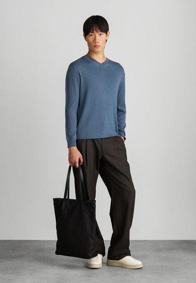 Ανοιχτό μπλε ριχτό πλεκτό πουλόβερ, χαλαρή εφαρμογή, V-λαιμόκοψη, συνδυασμένο με σκούρους καφετί παντελόνες και μαύρη τσάντα tote. Τα λευκά αθλητικά παπούτσια ολοκληρώνουν την εμφάνιση.