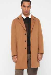 Uomo in un cappotto beige con bottoni sopra un maglione nero e una camicia bianca con colletto, che indossa pantaloni marroni su uno sfondo chiaro e semplice.