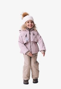 Veste isolante rose clair avec un accent en étoile, associée à un pantalon de neige beige et des bottes noires. Bonnet en tricot blanc avec un pompon marron sur le dessus.
