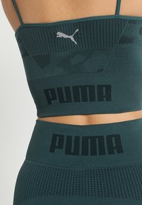 Tmavě zelená sportovní podprsenka s texturovanými síťovanými panely, s logem Puma a výrazným textem "PUMA" podél pásku. Stretchy, přiléhavý design.