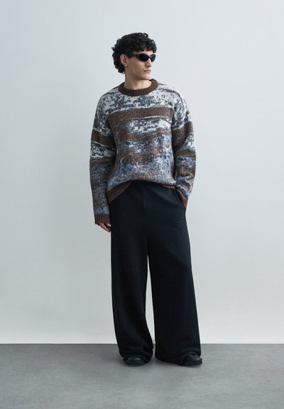 Knit pulover v rjavih, modrih in sivih odtenkih z okrolim izrezom in teksturiranim vzorcem. Kombiniran z ohlapnimi črnimi hlačami in črnimi čevlji.