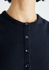 Haut noir à côtes avec une encolure arrondie, comportant six boutons ronds et foncés sur le devant pour la fermeture. La texture est douce et ajustée.