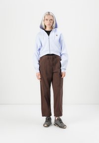 Sudadera recortada de cremallera color azul claro con un bolsillo frontal. El conjunto se completa con unos pantalones marrones de cintura elástica y zapatillas grises.