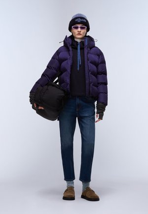 Lila puffjacka med hög krage, svart väska, denimjeans och bruna skor. Tillbehör inkluderar en stickad mössa och solglasögon.