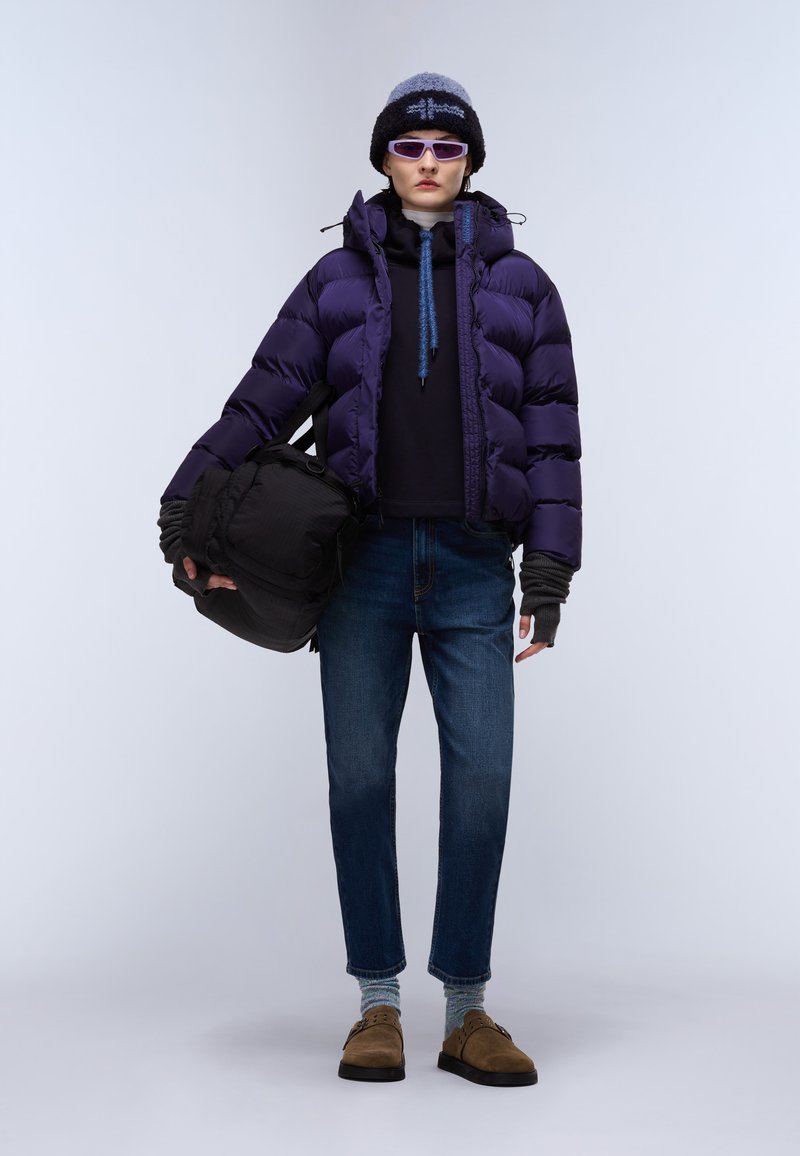 Fioletowa puchowa kurtka z wysokim kołnierzem, czarna torba, dżinsy i brązowe buty. Akcesoria to dziana czapka i okulary przeciwsłoneczne.