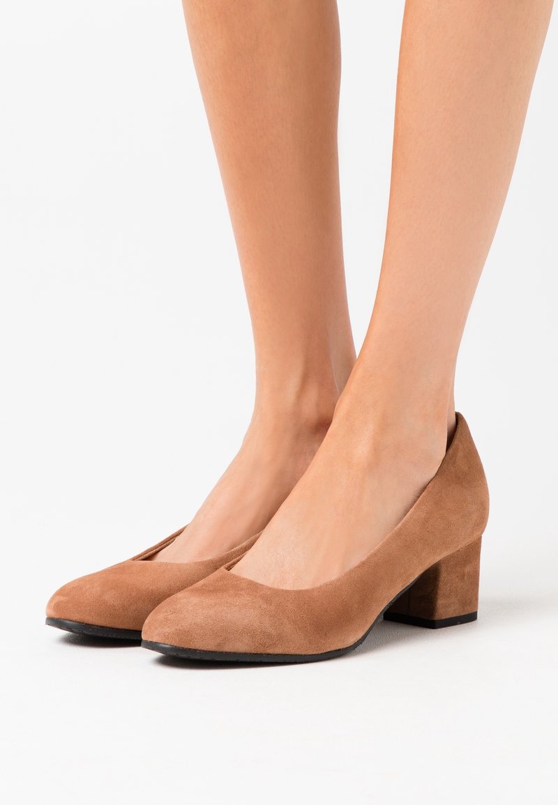 Des chaussures à talons blocs en suede de teinte neutre portées pieds nus, photographiées sur fond blanc.