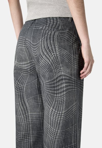 Primer plano de una persona que lleva pantalones anchos con patrón gris y negro, con detalle de bolsillo trasero, y una camiseta sin mangas de color gris claro.