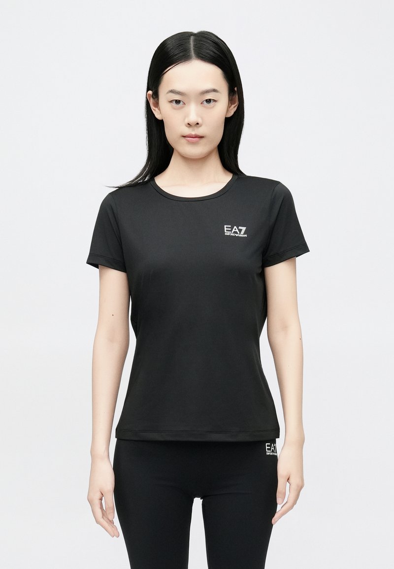 Fekete rövid ujjú póló sima anyagból, kerek nyakkivágással és egy kis fehér logóval a mellkason. Testhezálló szabás.