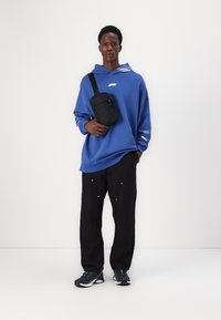 Modrá oversize mikina s bielym logom, čierna crossbody taška, čierne nohavice so striebornými akcentmi a tmavé športové topánky. Hladká textúra látky.